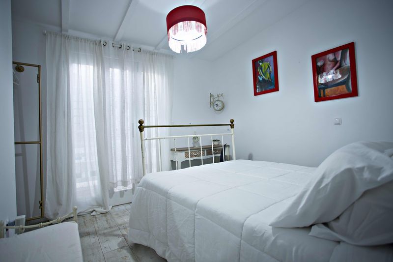 Cama de ferro branca junto a cortinas leves, com candeeiro vermelho e quadros